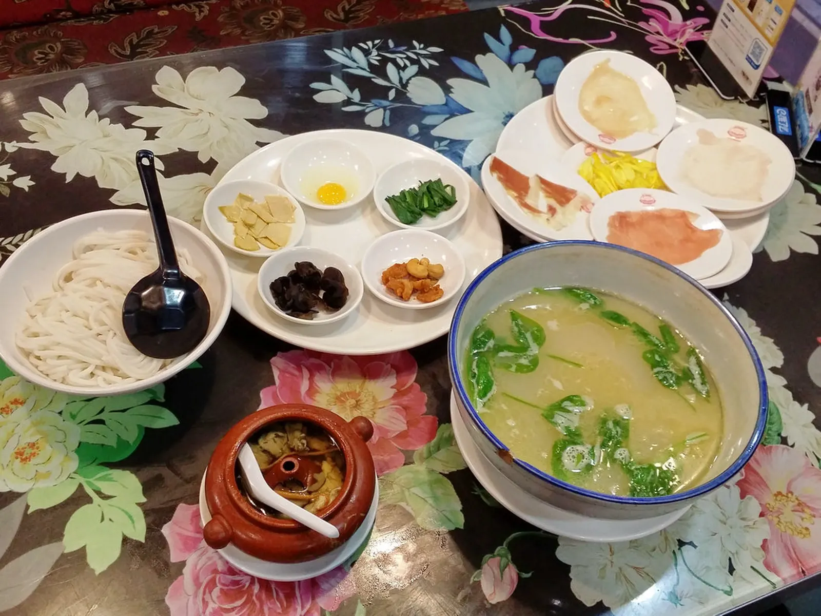 A crossing-the-bridge noodle set: raw plates arranged around a bowl of broth on a floral tablecloth.