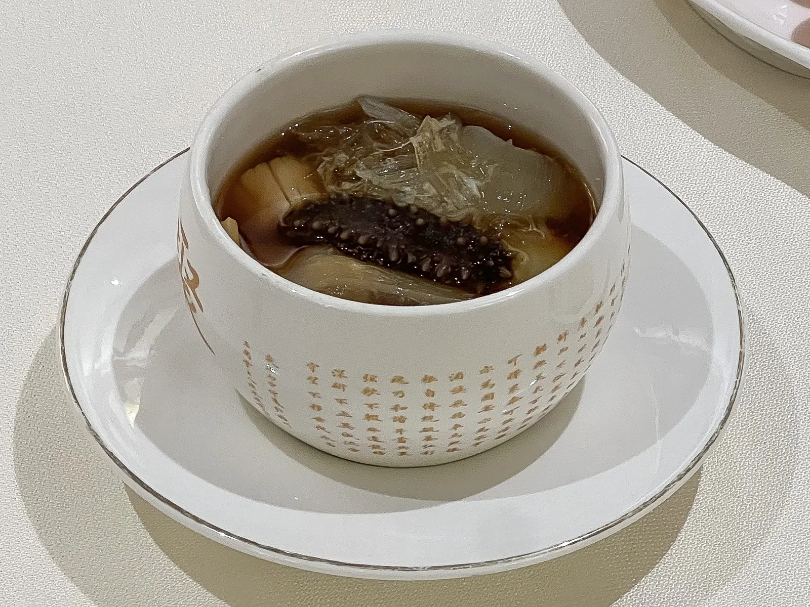 A single-serving pot of fotiaoqiang in printed porcelain, sea cucumber visible in the amber broth.