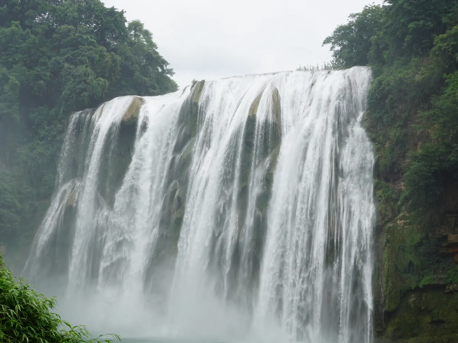 The full curtain of Huangguoshu Waterfall dropping into its green karst pool, framed by forest.