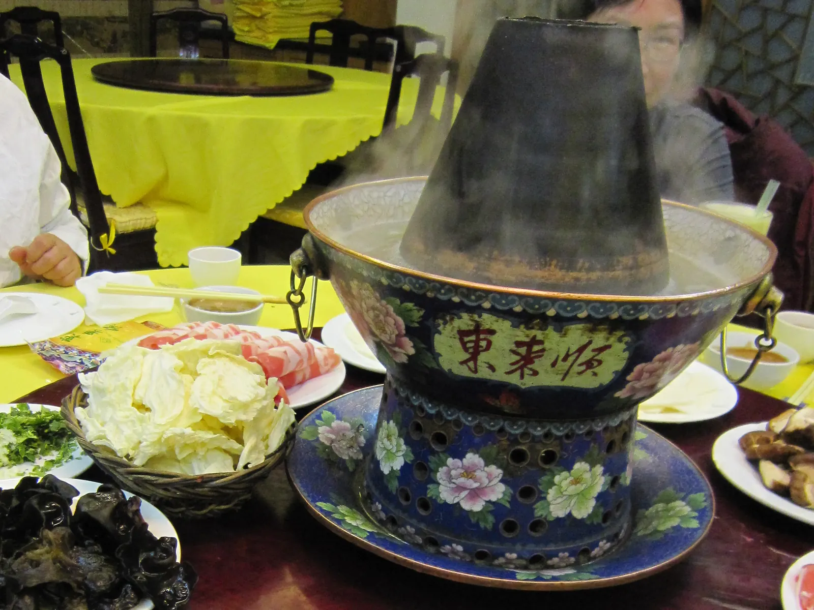 A charcoal copper hotpot for instant-boiled mutton, surrounded by plates of raw lamb rolls and cabbage.