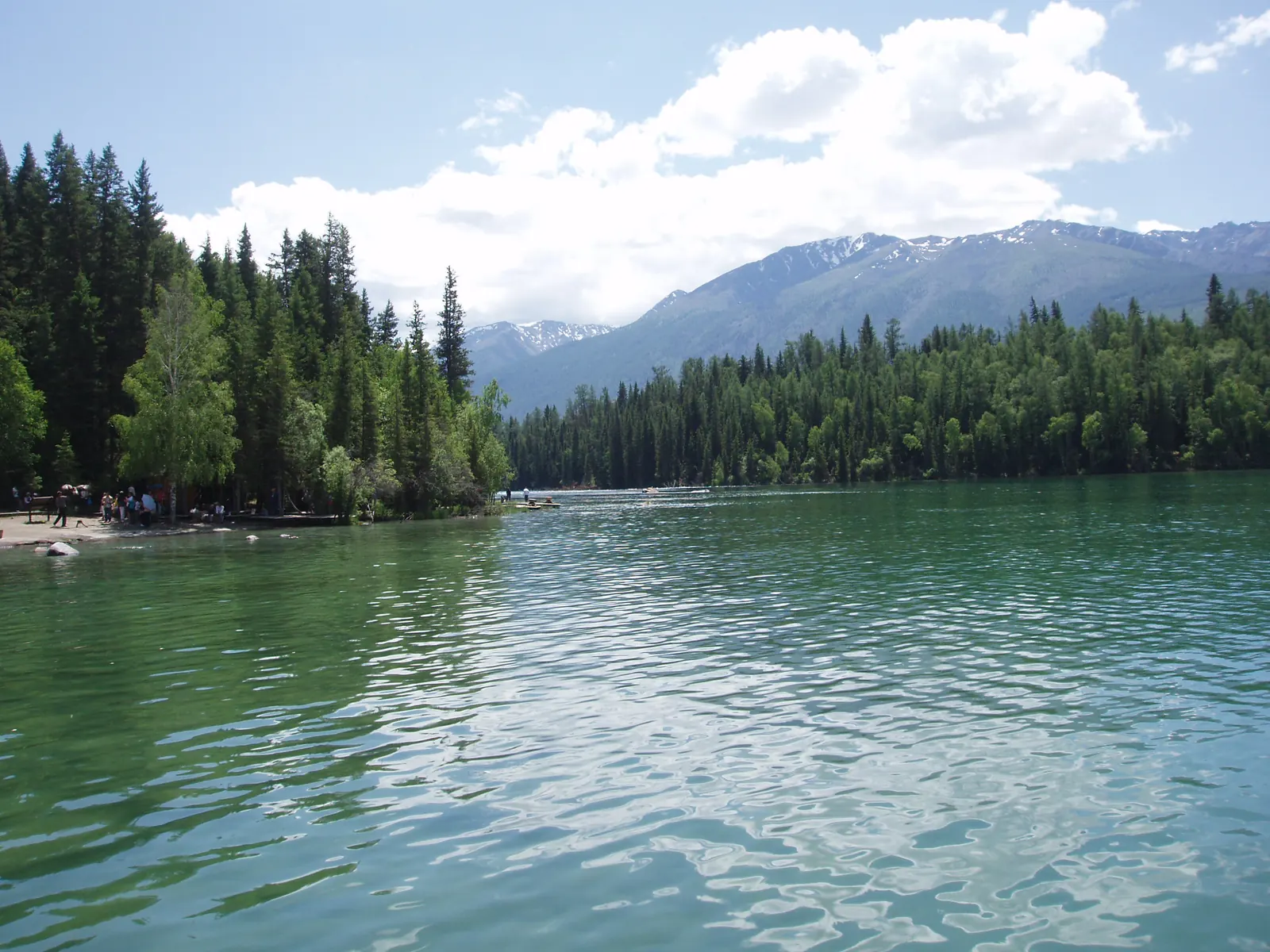 Kanas Lake framed by dark larch forest and snow-dusted mountains under a summer sky.
