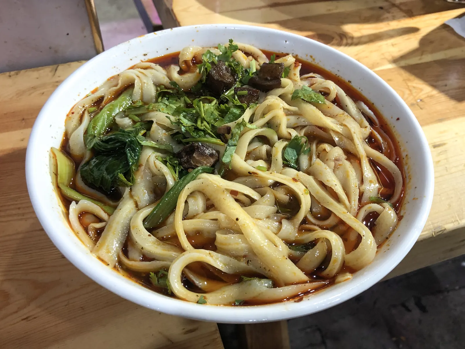 A bowl of knife-cut noodles in red broth with greens, shaved leaves thick in the middle and thin at the edge.