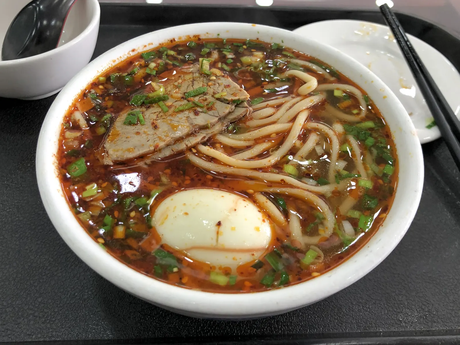 A bowl of Lanzhou beef noodle soup: red chili oil, hand-pulled noodles, beef slices, and an egg in clear broth.