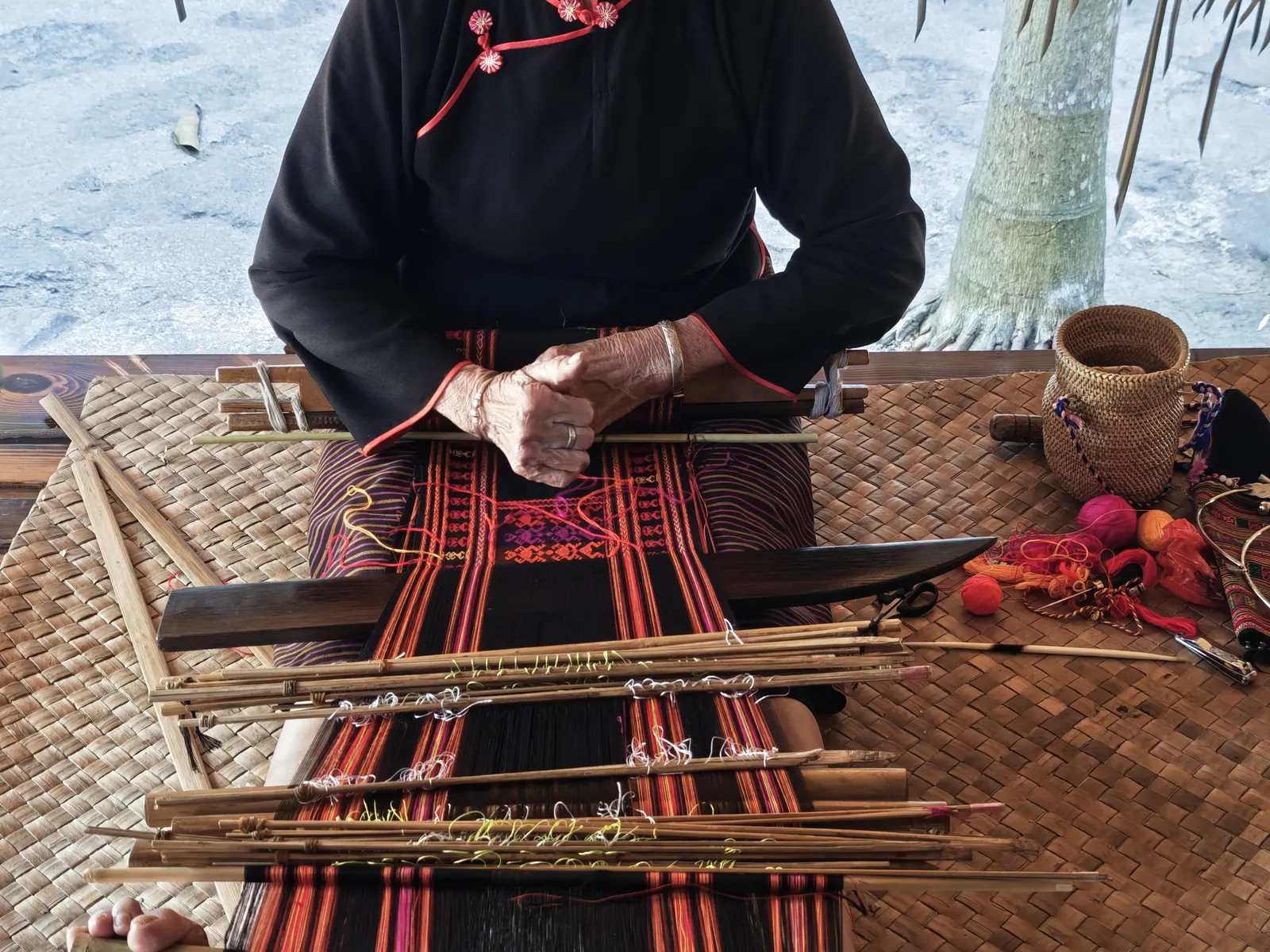 A Li weaver working a back-strap hip loom, brocade bands stretched before her on bamboo sheds.