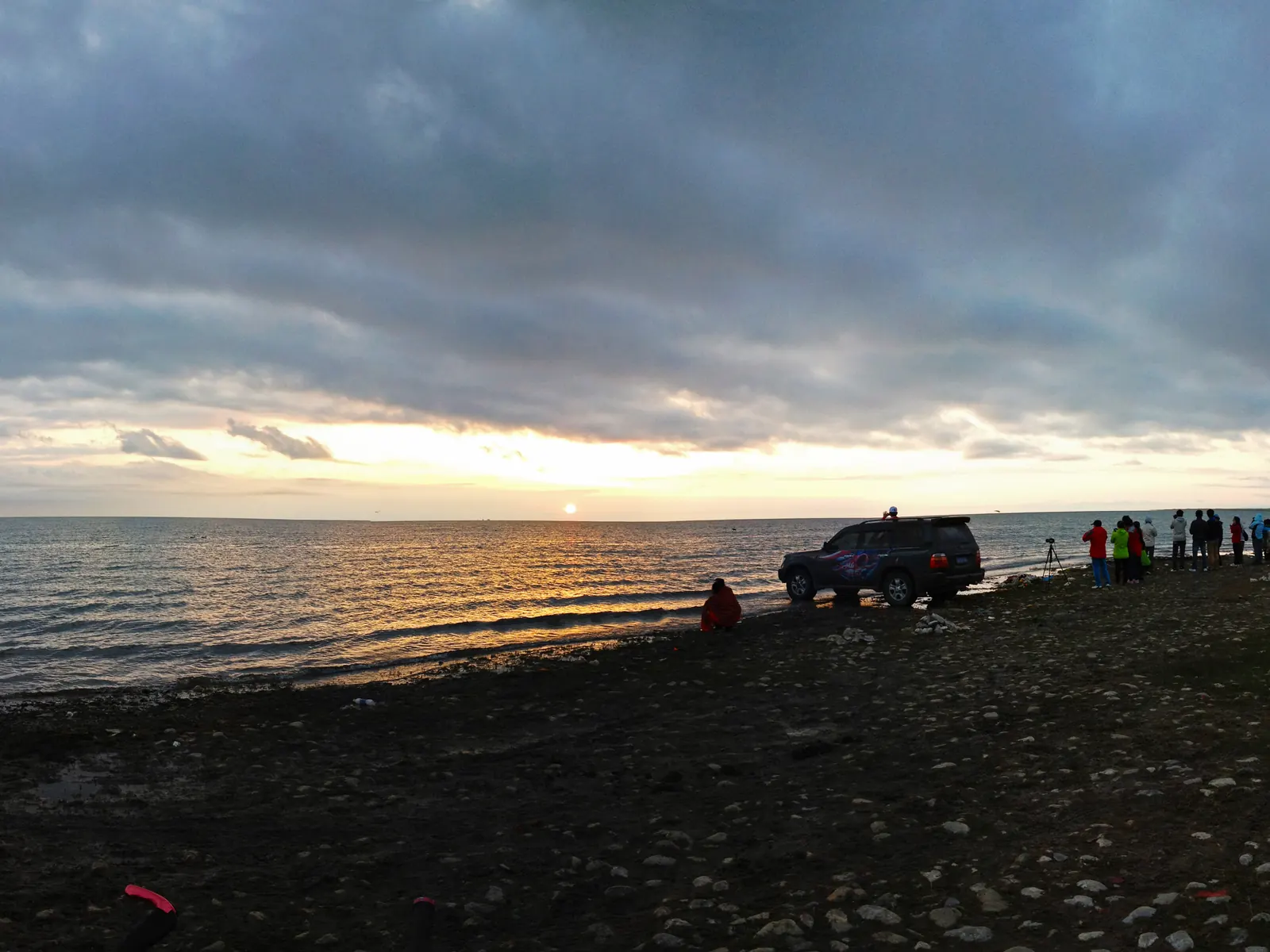 Sunrise over the shore of Qinghai Lake at Heimahe: grey dawn water, grassland, and a few early figures on the beach.