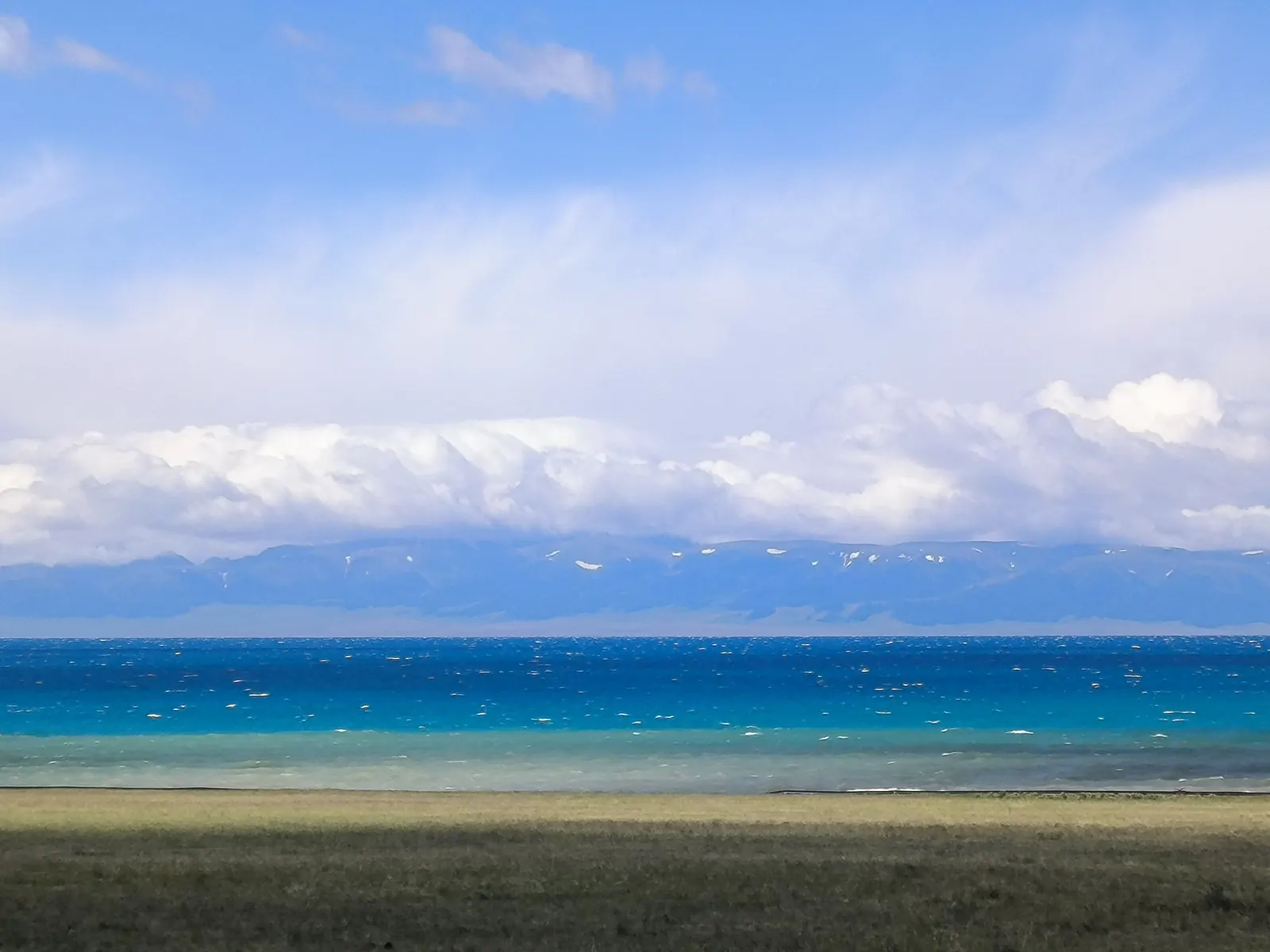 The deep layered blues of Sayram Lake under a wide sky, snow ridges on the far shore.