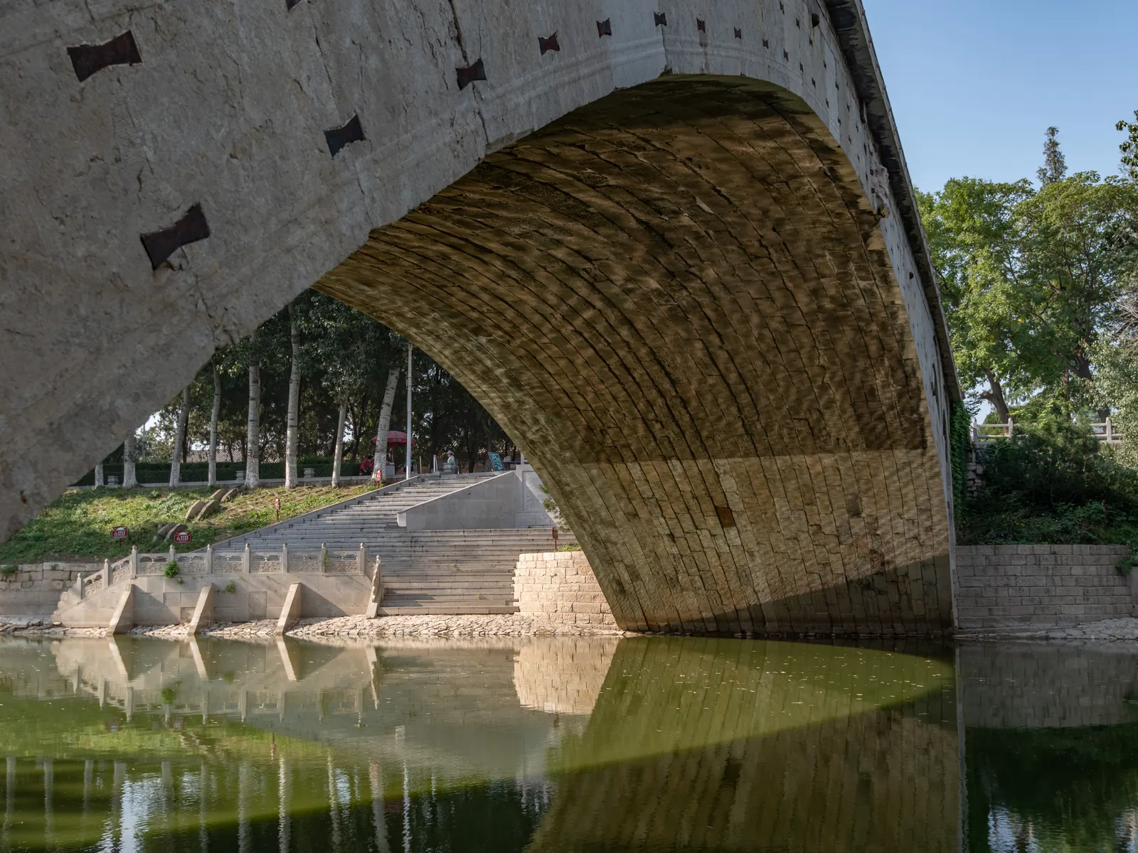 The great stone arch of Anji Bridge reflected in still water, wedge joints and iron stitches visible.